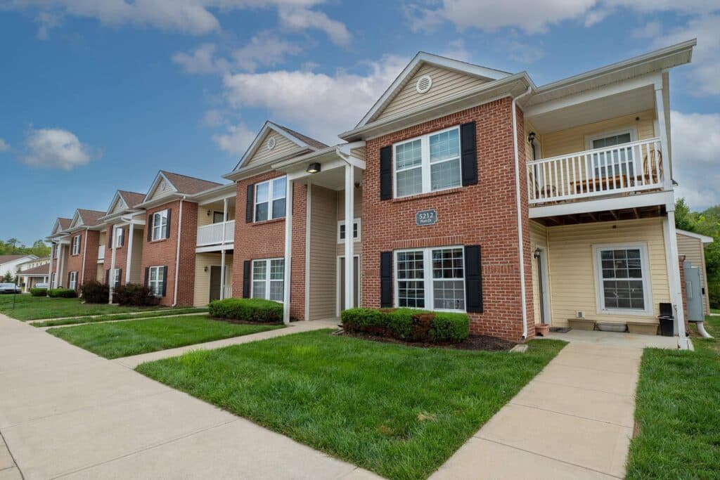 A row of two-story brick apartment buildings with white trim, black shutters, small balconies, and neatly maintained lawns under a partly cloudy sky.