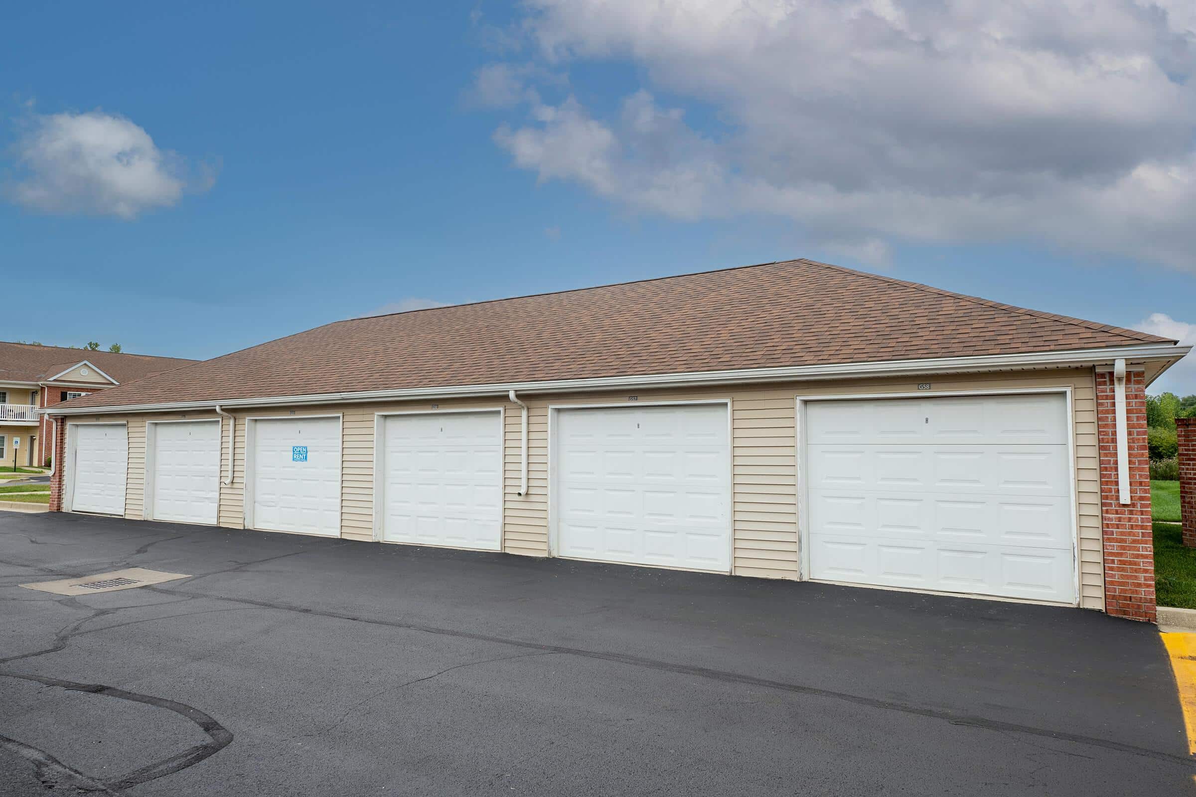 A row of six white garage doors in a single-story building with a brown shingle roof, next to a paved driveway under a partly cloudy sky.