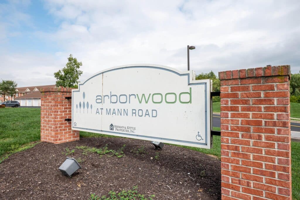 A large white sign reading "arborwood AT MANN ROAD" is displayed between two brick pillars on a landscaped area near a street.