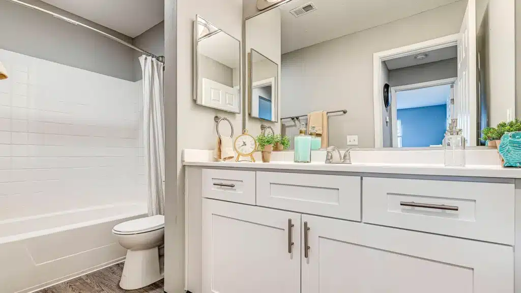 Modern bathroom with white cabinets, a countertop decorated with a clock and plants, a large mirror, and a bathtub-shower combination with a white curtain.
