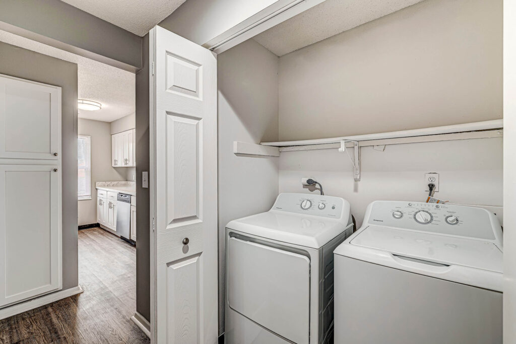 A laundry nook with a washing machine and dryer, open folding door, shelf above, and a view into a kitchen with white cabinets and wood flooring.