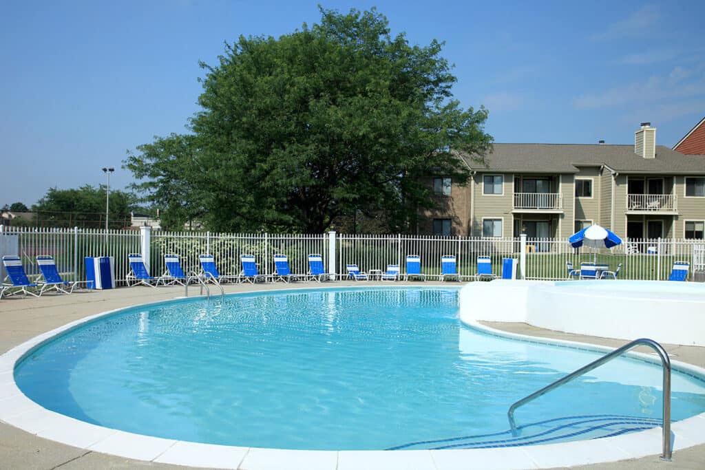 Outdoor swimming pool with clear water, surrounded by blue and white lounge chairs, tables, and umbrellas; apartment buildings and a large tree in the background.