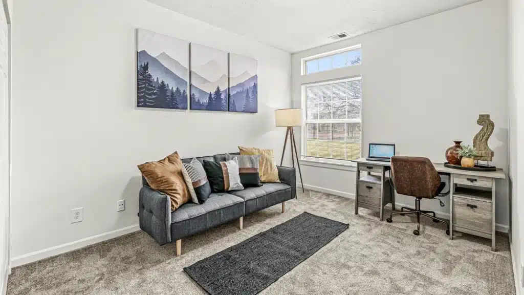 A bright home office with a gray sofa, decorative pillows, a floor lamp, wall art, a rug, and a desk with a laptop near a window.