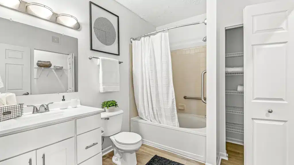 A white bathroom with a vanity, sink, toilet, potted plant, bathtub with a shower curtain, wall art, towel rack, and an open closet with shelves.