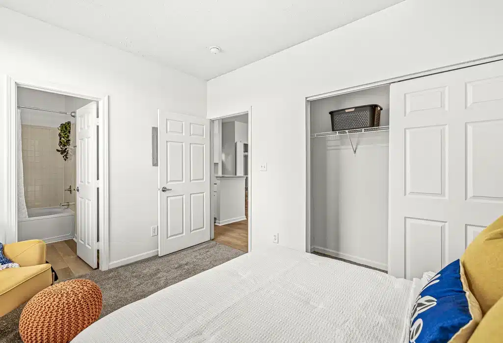 Bedroom with a bed, open closet, and doorways leading to a bathroom and hallway; includes a yellow chair and neutral decor.