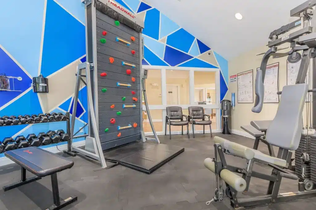 A gym room with a climbing wall, dumbbells, bench, weight machine, two chairs, water cooler, and geometric blue wall decor.