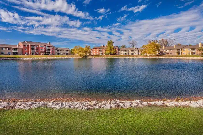Apartment buildings surround a pond under a blue sky with scattered clouds, bordered by a grass lawn and a strip of rocks in the foreground.