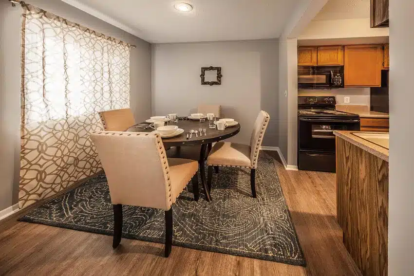 A dining area with a round table set for four, beige upholstered chairs, patterned rug, and an adjacent kitchen with wooden cabinets and black appliances.
