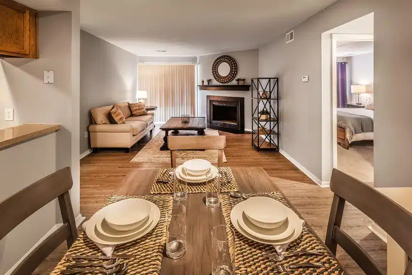 Dining table set for four in the foreground, with a view of a living room featuring a sofa, coffee table, fireplace, shelving unit, and an adjacent bedroom.