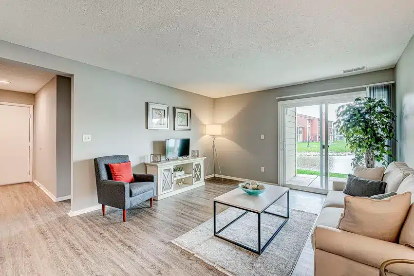 Modern living room with neutral decor, gray armchair, beige sofa, coffee table, TV stand, floor lamp, and sliding glass doors leading to an outdoor area.