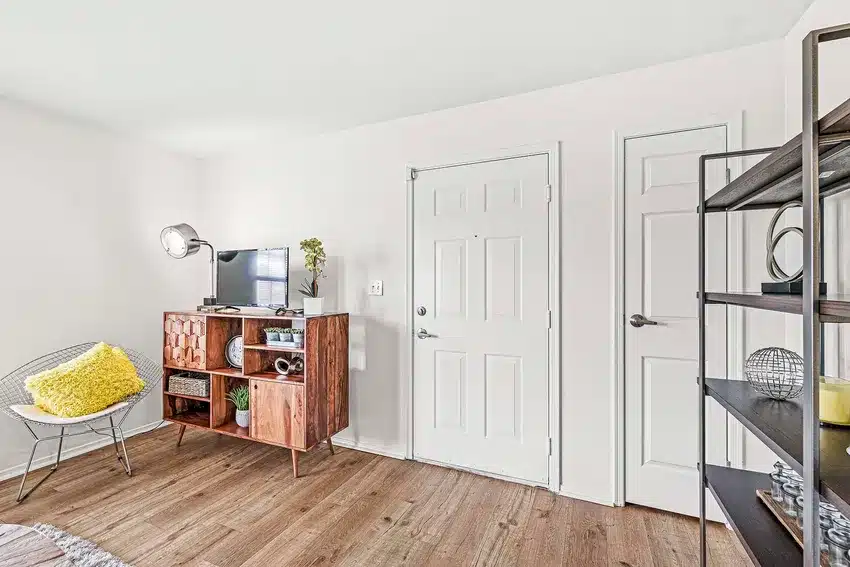 A room with wood flooring, a mid-century style TV stand, a chair with a yellow pillow, two white doors, and a metal shelving unit with decorative items.