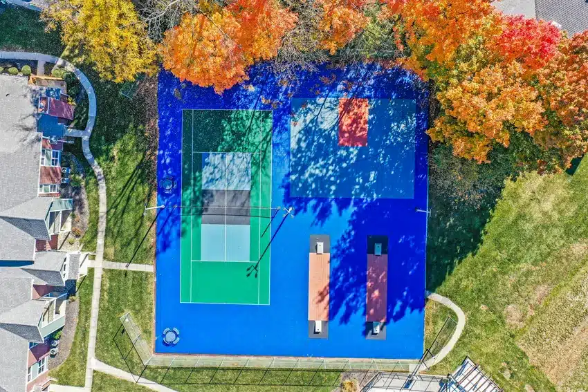Aerial view of a blue multi-use sports court with tennis and basketball markings, surrounded by autumn trees and nearby residential buildings.