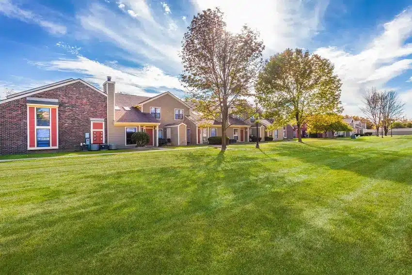 Row of suburban houses with brick and siding exteriors, trees, and a large, well-maintained grassy lawn under a blue sky with clouds.