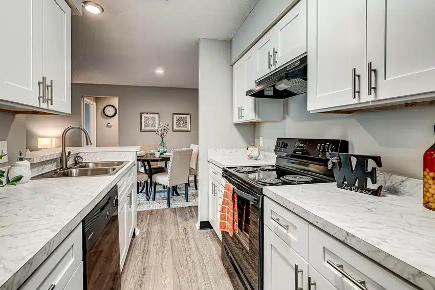Modern galley kitchen with white cabinets, marble countertops, black appliances, and a view into a dining area with a table and upholstered chairs.
