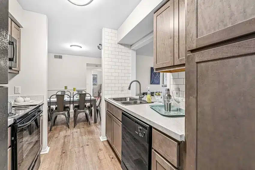 A modern galley kitchen with wood cabinets, stainless steel sink, dishwasher, electric stove, and view into a dining area with metal chairs and a set dining table.