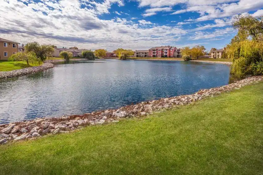 A calm pond surrounded by rocks and grass, with apartment buildings and trees in the background under a partly cloudy sky.