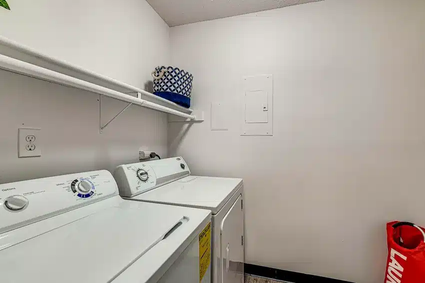 A laundry room with a white washing machine and dryer, a wall shelf holding baskets, and a red laundry bag on the floor.