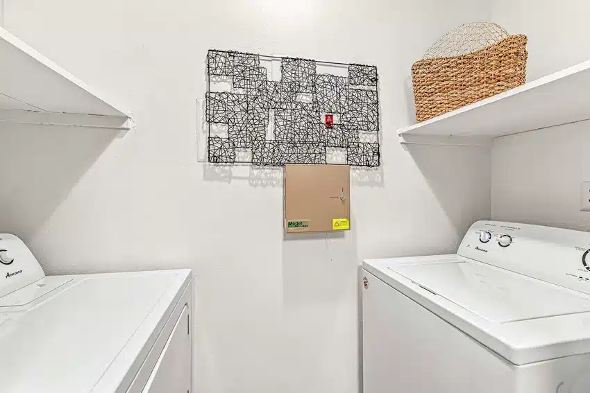 Laundry room with a white washer and dryer, white walls, open shelves, a brown basket on the right, and electrical panel covers on the back wall.