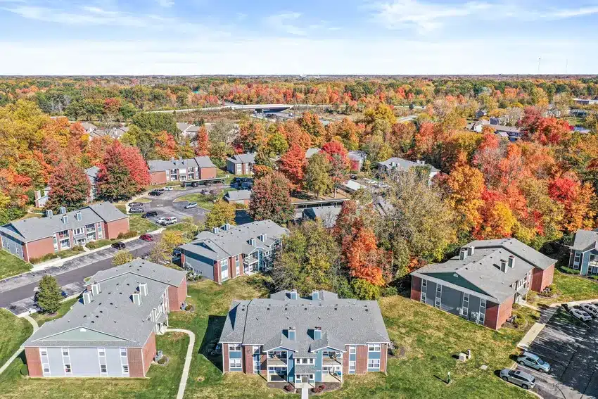 Aerial view of a suburban apartment complex surrounded by colorful autumn trees, with roads and parking lots visible among the buildings.