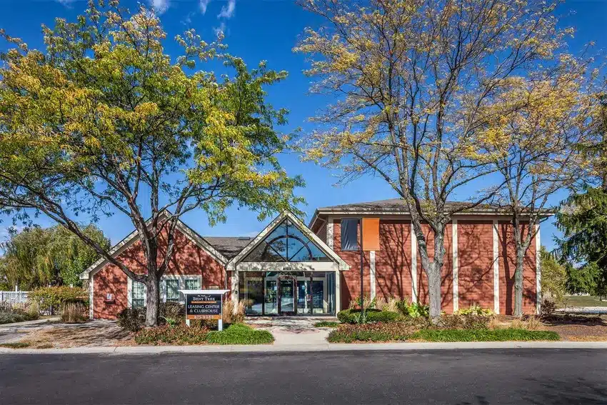 A brick building with large windows and a peaked glass entrance sits behind trees with autumn foliage, under a clear blue sky.