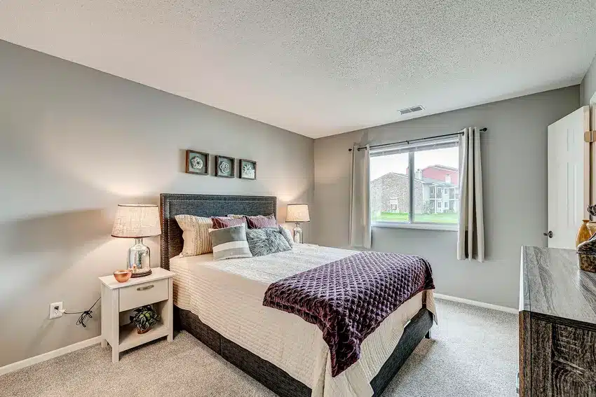 A neatly made bed with pillows and a purple throw blanket is centered in a carpeted bedroom with gray walls, two nightstands, lamps, and a dresser by a window with curtains.