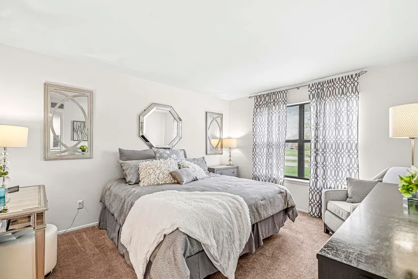 A neatly made bed with gray and white bedding is centered in a bright bedroom with two windows, bedside tables, lamps, and decorative mirrors on the walls.
