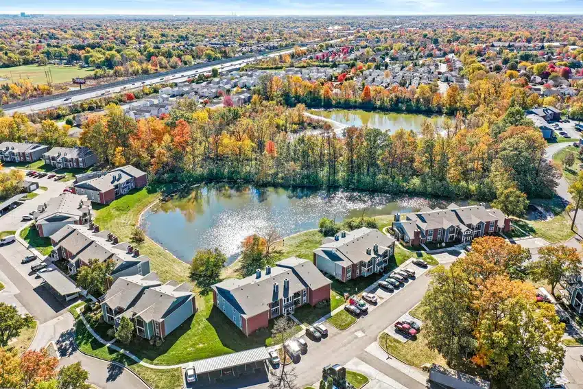 Aerial view of a suburban residential area with apartment buildings, parking lots, trees with autumn foliage, and a small lake in the center.