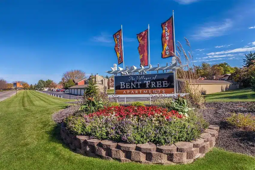 A landscaped flower bed surrounds a sign reading "The Villages of Bent Tree Apartments" with three vertical flags behind it, along a suburban street.