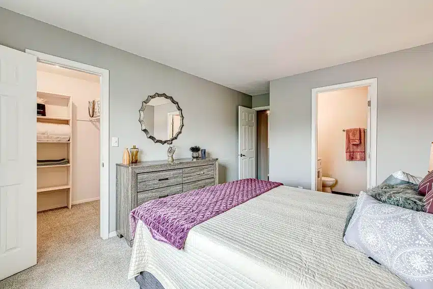 Bedroom with a neatly made bed, dresser with mirror, open doors showing a walk-in closet and a bathroom, both with towels and shelves visible.