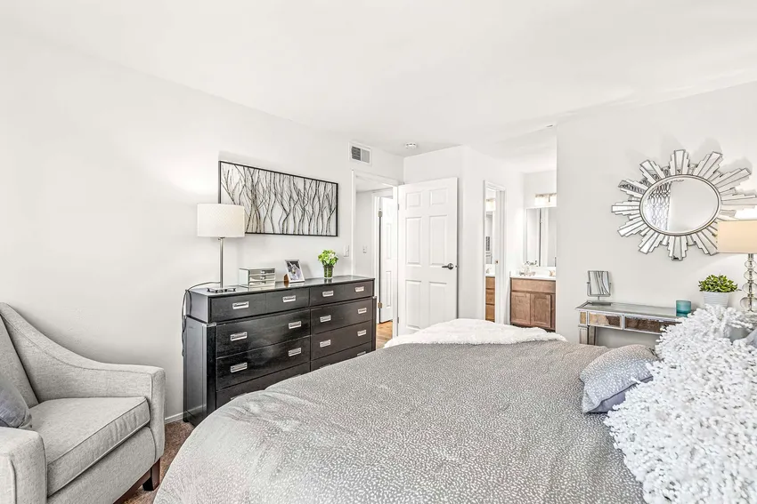 A modern bedroom with a neatly made bed, gray dresser, armchair, wall art, and a sunburst mirror above a console table; a bathroom is visible through an open door.