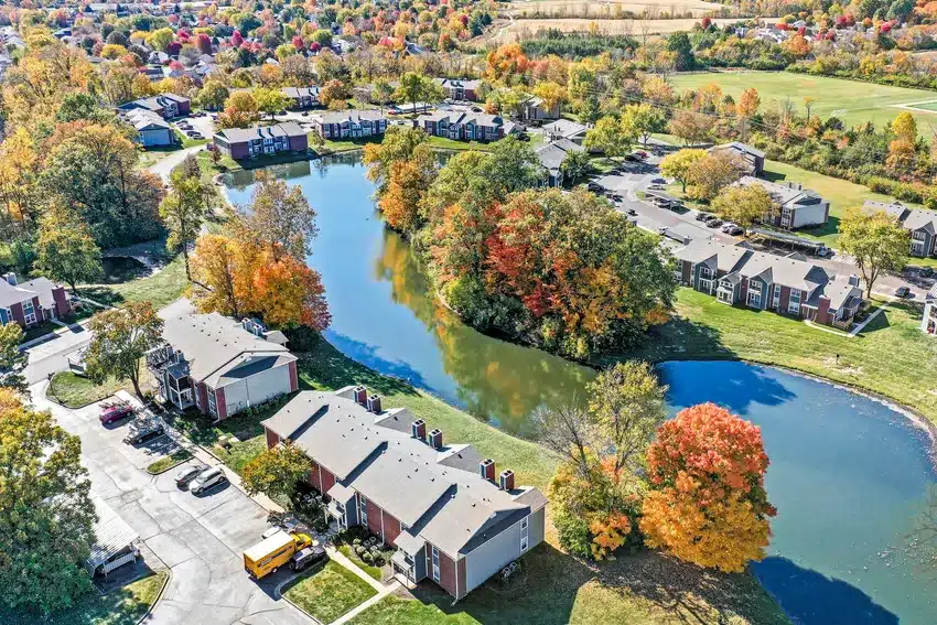 Aerial view of an apartment complex with several buildings, parking lots, a pond, and surrounding trees with fall foliage.