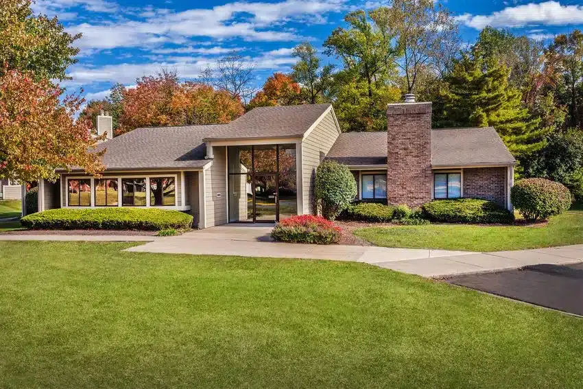 A single-story house with large windows, a brick chimney, and a covered entryway, surrounded by green lawn, shrubs, and trees with autumn foliage.