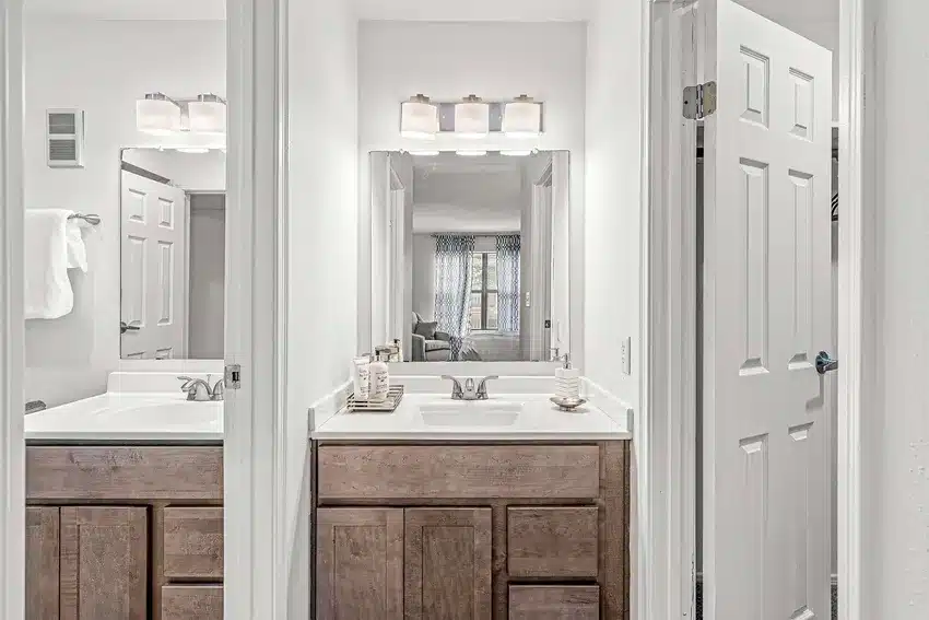 A modern bathroom with a double sink vanity, wooden cabinets, large mirrors, and bright lighting; a towel hangs on the wall and an open door reveals a glimpse of a bedroom.