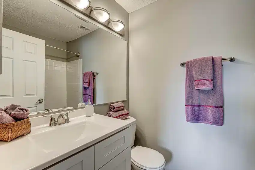 Modern bathroom with a large mirror, white vanity, silver faucet, toilet, towel rack with purple towels, and a basket of towels on the counter.