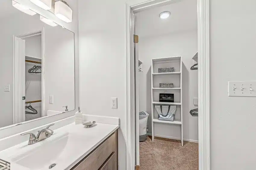 A bathroom with a sink and mirror in the foreground, and an open door revealing a walk-in closet with shelves and hanging space in the background.