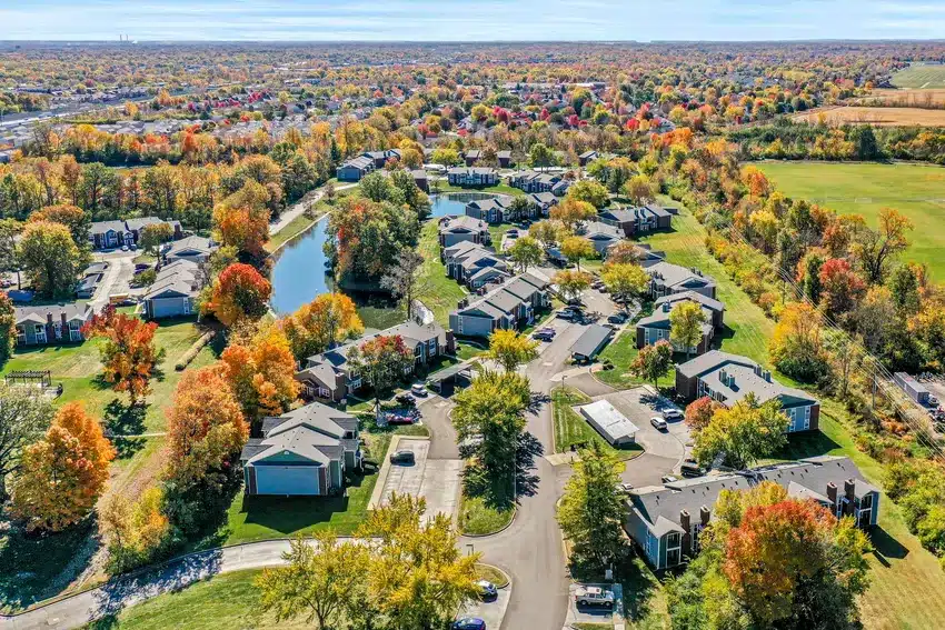 Aerial view of a suburban neighborhood with houses, roads, and a pond, surrounded by colorful autumn trees and open green fields.