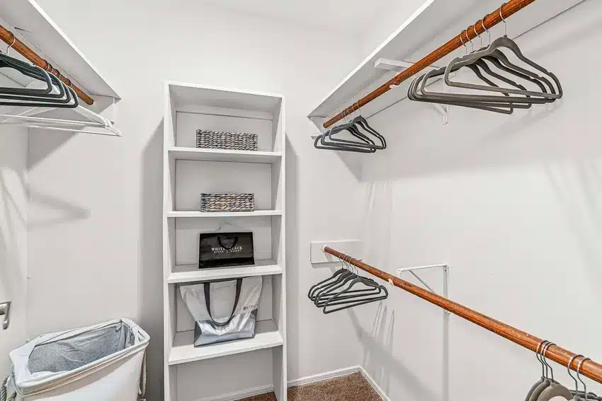 Walk-in closet with white shelves, wooden hanging rods, black hangers, two baskets, a laundry bin, and a brown carpeted floor.