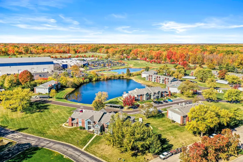 Aerial view of a residential community with apartments, a pond, green lawns, trees with autumn foliage, and clear blue sky.