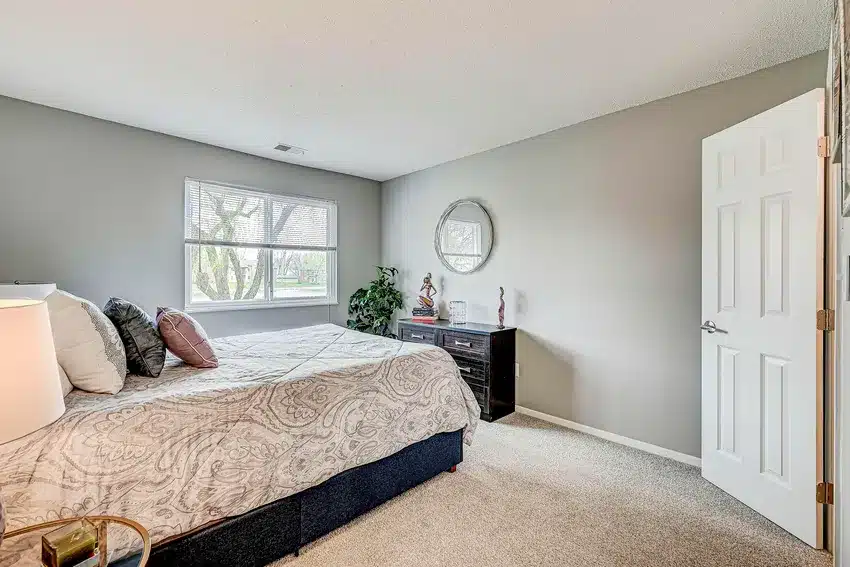 A bedroom with a bed, patterned bedding, two pillows, a dresser with decor, a round wall mirror, a potted plant, and a window letting in daylight.
