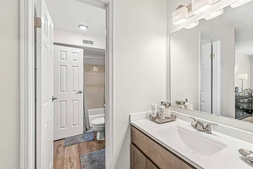 Modern bathroom with a single-sink vanity, rectangular mirror, and neutral decor. Toilet and shower area visible in the background with white doors and wood-look flooring.