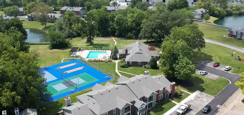 Aerial view of an apartment complex featuring a swimming pool, sports courts, parking lot, green lawns, and surrounding trees.