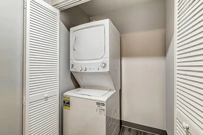Stacked washer and dryer unit in a small closet with white louvered doors, set against light-colored walls and a dark floor.