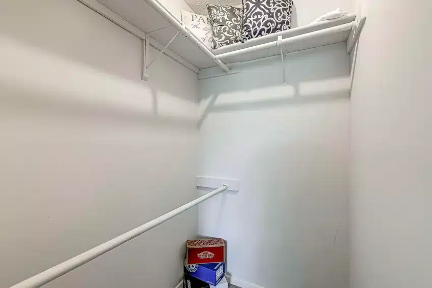 A small walk-in closet with white walls and shelves, a few decorative pillows on the top shelf, and two boxes on the floor below a hanging rod.