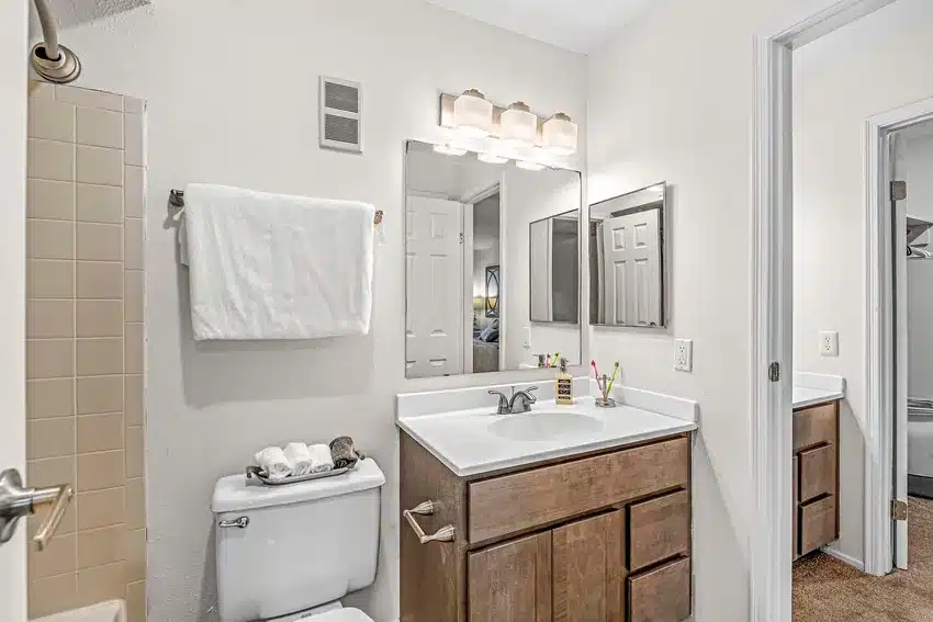 Modern bathroom with a large mirror, wooden vanity, white countertop, towel rack above the toilet, and tiled shower partially visible on the left.