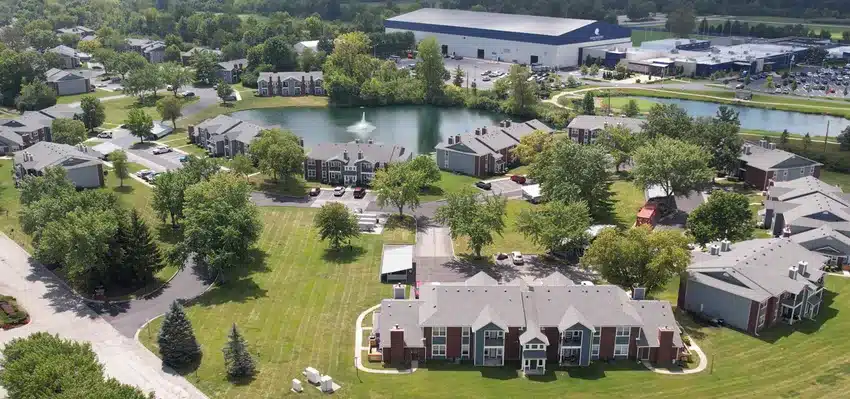 Aerial view of an apartment complex with multiple buildings, green lawns, trees, a central pond with a fountain, and a large industrial building in the background.