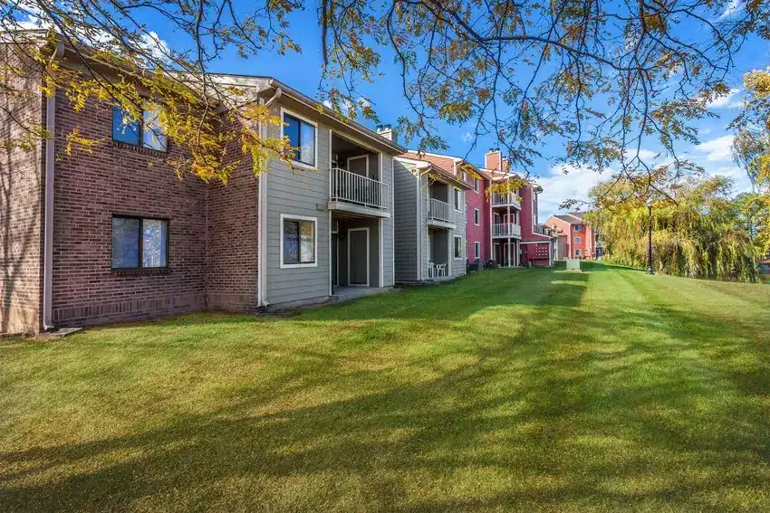 A row of apartment buildings with patios and balconies overlooks a grassy lawn on a sunny day, with trees casting shadows on the grass.