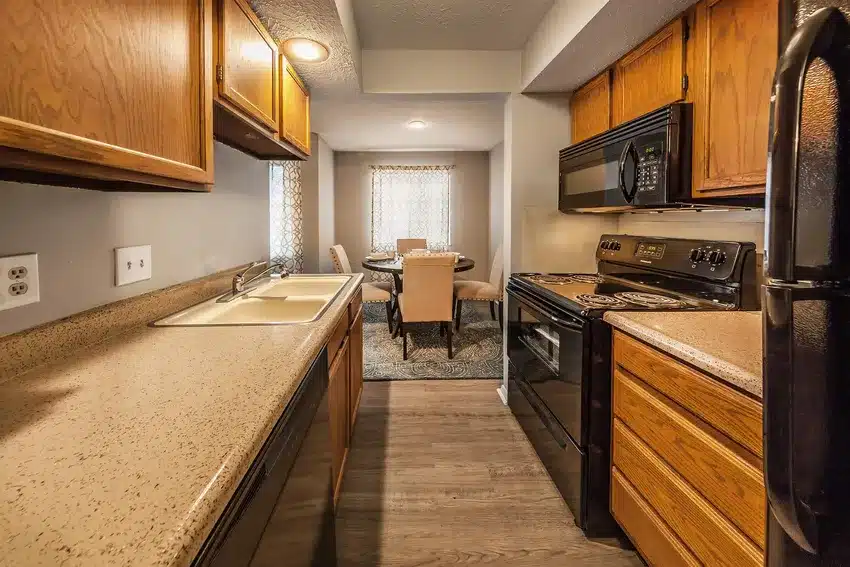 Galley-style kitchen with wood cabinets, laminate countertops, black appliances, and a dining area with a table and chairs visible in the background.
