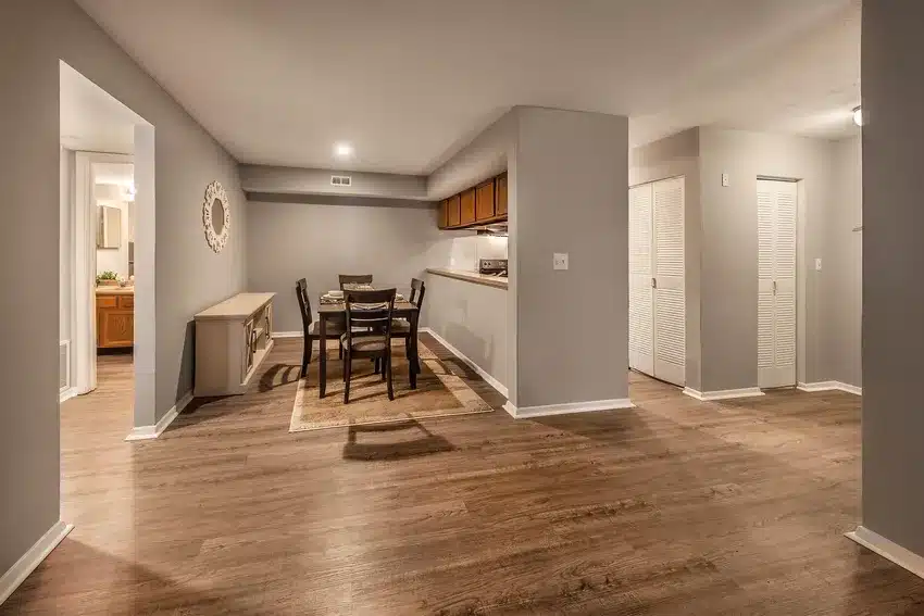 Open dining area with a table and four chairs, wood flooring, neutral walls, and adjacent kitchen and hallway with white closet doors.
