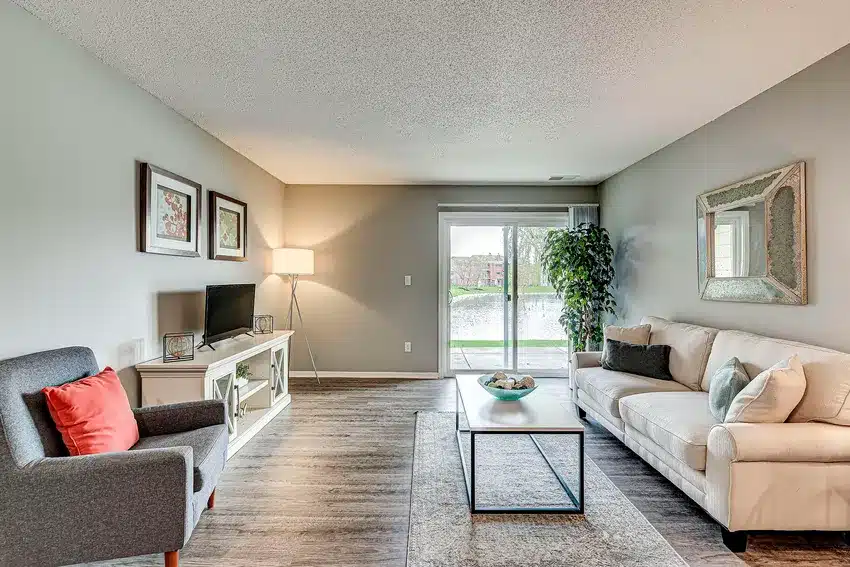 A modern living room with a gray armchair, beige sofa, coffee table, TV stand, and large window overlooking a patio with greenery outside.