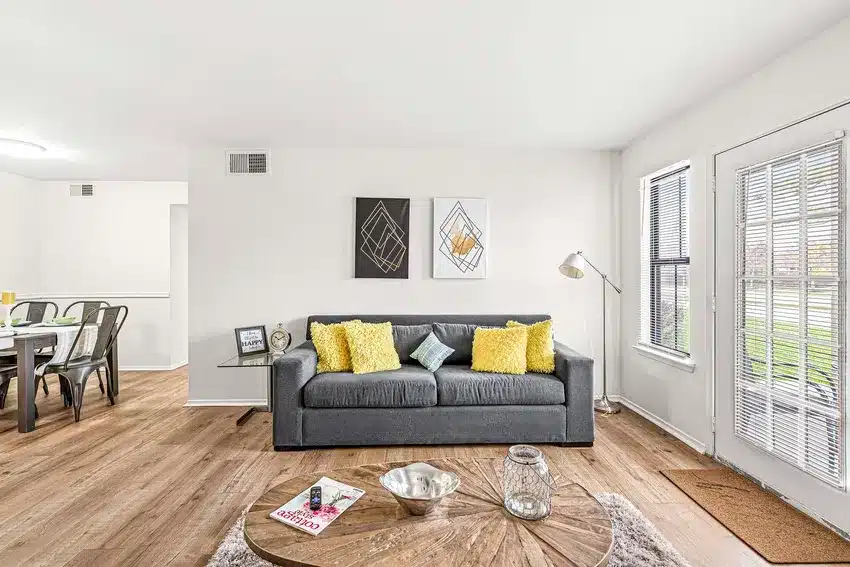 Modern living room with a gray sofa, yellow cushions, minimalist wall art, and a round wooden coffee table. Large windows and a door let in natural light. Dining area visible in the background.